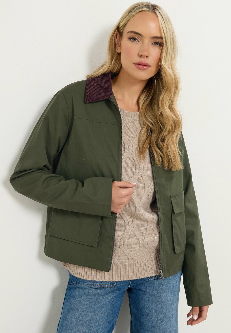 Oliven grøn jakke med en kordfløjl burgunder krave, lynlås foran og store lommer. Båret over en beige kabelstrikket sweater og blå jeans.