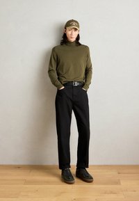 Dolcevita verde oliva, pantaloni neri e sneakers olive con suole marroni. Il modello indossa un cappellino abbinato con dettagli del logo.
