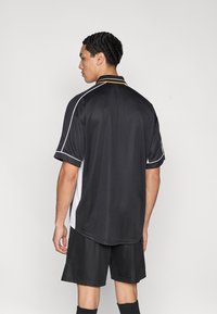 Μαύρο αθλητικό πόλο με λευκές και πορτοκαλί λεπτομέρειες, κοντά μανίκια και υφή σε φινίρισμα. Φοριέται με μαύρο σορτς.