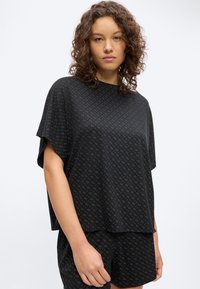 Γυναίκα με σγουρά μαλλιά, φορώντας ένα φαρδύ μαύρο τοπ με μοτίβο και κοντά παντελονάκια που ταιριάζουν, στέκεται μπροστά σε λευκό φόντο.