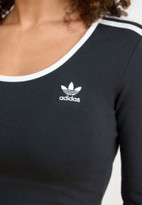 Musta pitkähihainen paita, jossa on ribbikangasta, valkoinen pyöreä pääntie ja pieni valkoinen Adidas-logo rinnassa.