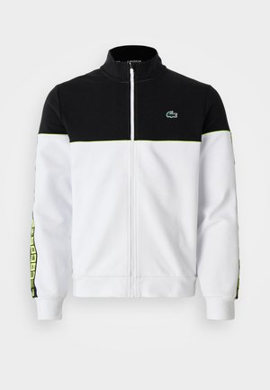 Giacca con zip completa, parte superiore nera, parte inferiore bianca, rifiniture verde fluorescente, strisce laterali a motivi e piccolo logo di coccodrillo verde sul petto.