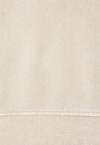 Béžová bavlněná tkanina s měkkou texturou. Na spodním okraji je viditelný šev, který vytváří jemný kontrast v odstínech.