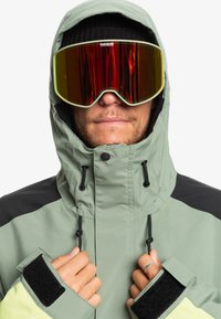 Lys grøn vandtæt jakke med sorte accenter, høj krave og justerbare manchetter. Store spejlede skibriller med rød toning.