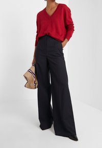 Pull à col en V rouge associé à un pantalon large à rayures bleu marine. Le mannequin tient un sac beige avec un accent à rayures foncées, portant des chaussures noires à bout pointu.