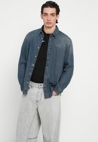 Πουκάμισο από denim σε σκούρο μπλε με κουμπιά και κεντητό λογότυπο, συνδυασμένο με ανοιχτό γκρι, φαρδιά τζιν παντελόνια με εμφανείς ραφές.