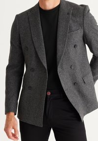 Grijze wollen dubbelgebreide blazer met klepkragen, voorzien van zwarte knopen en een borstzak, gedragen over een zwarte trui met ronde hals.