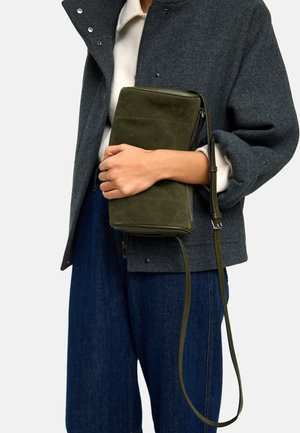 Osoba ubrana w ciemnoszarą kurtkę i niebieskie dżinsy trzyma oliwkowozieloną prostokątną torbę zamszową na ramię z długim paskiem.