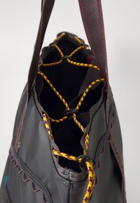 Sac en tissu gris avec une surface texturée. Doté d'une fermeture à cordon avec des cordons multicolores, des coutures rouges et un bord décoratif.