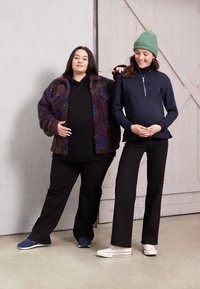Duas mulheres grávidas estão juntas. Uma delas usa uma jaqueta felpuda, colorida e com padrões, sobre um moletom preto e umas calças, enquanto a outra usa um top azul-marinho com fecho e umas calças pretas.