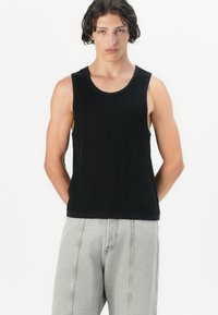 Sort ribbet tanktop med rund halsudskæring, parret med lysegrå denimbukser med synlige sømme. Modellen står med hænderne placeret bag ryggen.