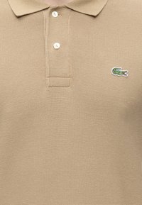 Polo beige texturé avec col, deux boutons blancs et un petit logo crocodile vert sur la poitrine.