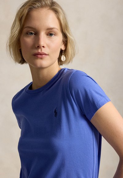 Ξανθιά γυναίκα με κοντά μαλλιά που φοράει μπλε μπλούζα με μικρό λογότυπο και ένα σκουλαρίκι με κοχύλι, κοιτάζει ήρεμα στην κάμερα.