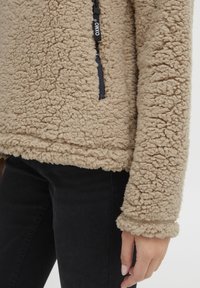 Pull-over en polaire beige avec un extérieur texturé et moelleux, comportant une fermeture éclair sur le côté et des poignets en maille côtelée. Le tissu est doux et épais.
