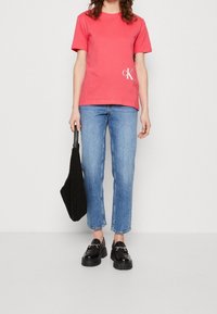 Korálové tričko s bílým logem "CK" na spodním lemu, v kombinaci se světle modrými džínami rovného střihu. Černá taška a robustní černé mokasíny dodávají vzhledu finální dotek.