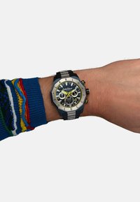Schwarze und silberne Armbanduhr mit achteckigem Design, ausgestattet mit einem schwarzen Zifferblatt, gelben Akzenten und silbernen Elementen, getragen an einer Hand mit einem blauen Pullover.