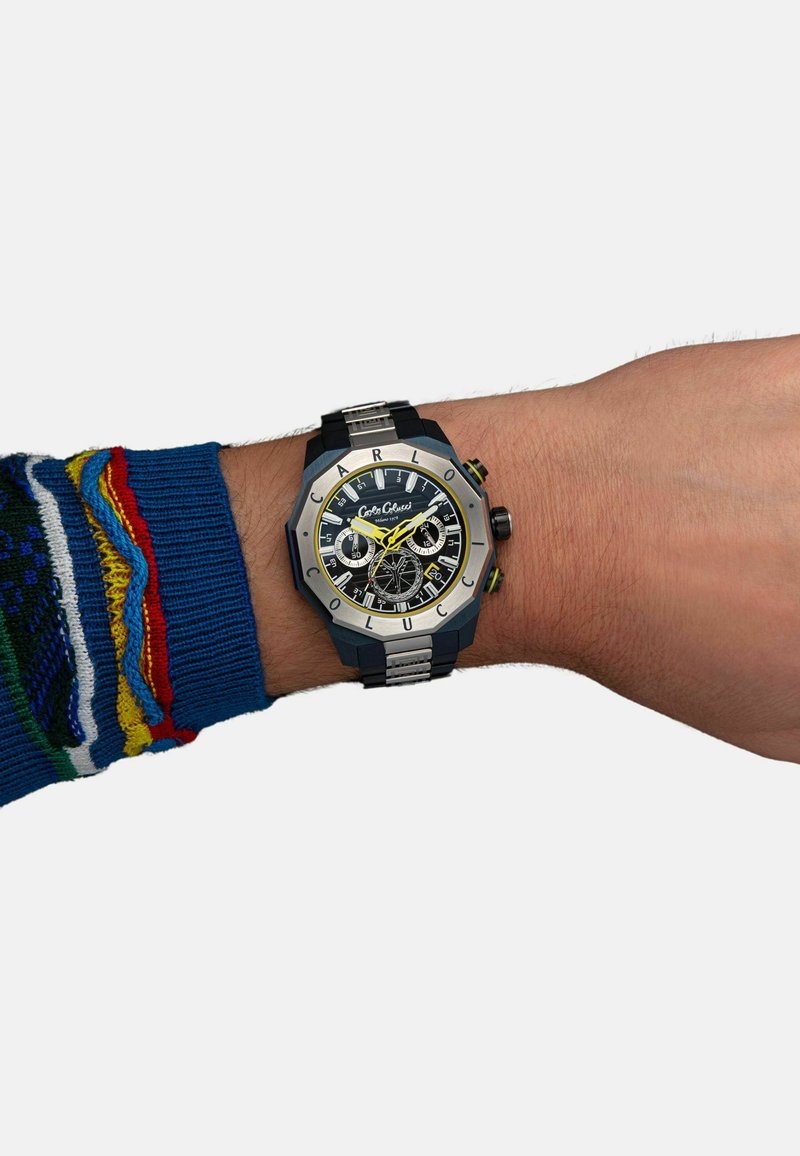 Schwarze und silberne Armbanduhr mit achteckigem Design, ausgestattet mit einem schwarzen Zifferblatt, gelben Akzenten und silbernen Elementen, getragen an einer Hand mit einem blauen Pullover.