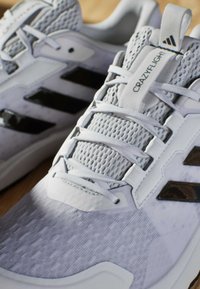 Szare buty sportowe z siateczkowymi i syntetycznymi materiałami, z trzema czarnymi paskami, teksturowanymi szarymi akcentami i białymi sznurówkami.