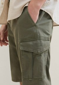 Pantalones cortos en verde oliva de estilo cargo, confeccionados con tejido texturizado, que presentan bolsillos laterales y un detalle cosido en la cinturilla y los bolsillos.