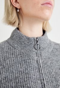 Szary sweter dzianinowy z wysokim kołnierzem i zamkiem błyskawicznym, posiadający okrągły uchwyt oraz prążkowaną strukturę. Zbliżenie podkreślające detale.