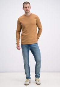 Brązowa bluza z długim rękawem i okrągłym dekoltem, z prążkowanymi mankietami i pasem, zestawiona z jasnoniebieskimi dżinsami i białymi sneakersami z beżowymi akcentami.