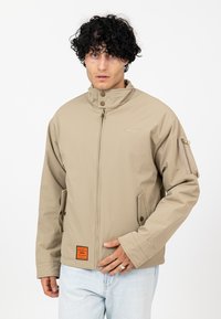 Beżowa wodoodporna kurtka z wysokim kołnierzem, z przodu na zamek, z kieszonką na piersi i pomarańczowym detalem, o gładkiej matowej teksturze.