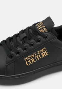 Svart lädersneaker med rund tå, guldlogotyp "VERSACE JEANS COUTURE", texturerade snören och en slät yta. Gummisulor för bra grepp.