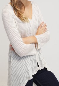 Lehký pruhovaný úpletový top s výstřihem do V, dlouhými rukávy a zaobleným lemem. Vyznačuje se měkkou, texturovanou látkou v krémové a světle béžové barvě.