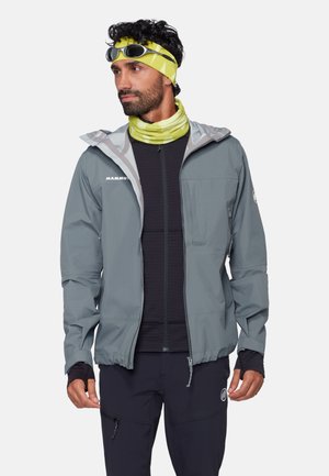 Mann trägt eine graue Outdoorjacke über einem schwarzen Reißverschlusspullover, schwarze Hose, gelb-grünes Stirnband und Nackenwärmer, mit Sonnenbrille auf dem Kopf.
