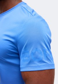 Κοντινό πλάνο του άνω βραχίονα ενός ατόμου που φορά ένα ανοιχτό μπλε, υφασμάτινο κοντομάνικο πουκάμισο με μερικό ανάγλυφο κείμενο στο μανίκι.