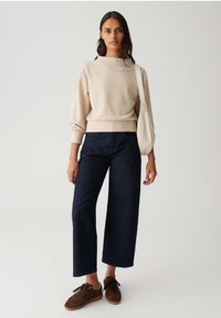 Felpa beige con scollo a barca e maniche arricciate, abbinata a jeans a gamba larga blu scuro e sneakers marroni. Outfit casual.