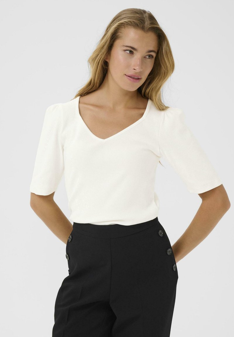 Top bianco a coste con scollatura a V e maniche a puff corte, abbinato a pantaloni neri a vita alta con bottoni decorativi sul lato.