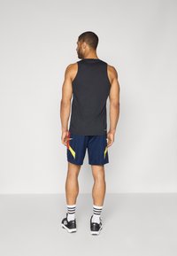 Zwarte mouwloze tanktop gecombineerd met marineblauwe shorts met gele accenten en een mesh-structuur. Zwarte sportschoenen met zwart-wit gestreepte sokken.