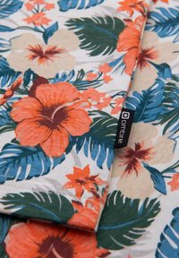 Bloemstof met oranje en beige hibiscus bloemen, groene bladeren en blauwe accenten. Inclusief een klein zwart label met de merknaam "ombre."