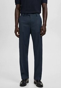 Hombre vestido con pantalones azul marino a medida, mocasines negros y una camisa oscura metida por dentro, de pie contra un fondo claro y liso.