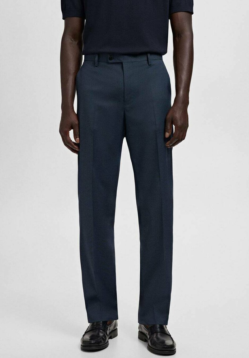 Hombre vestido con pantalones azul marino a medida, mocasines negros y una camisa oscura metida por dentro, de pie contra un fondo claro y liso.