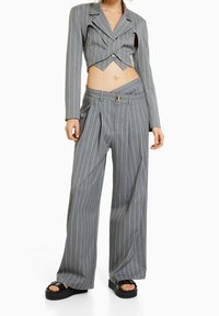 Vrouw in een grijze gestreepte cropped blazer en bijpassende wijde broek met een asymmetrische tailleband en zwarte plateau-sandalen.