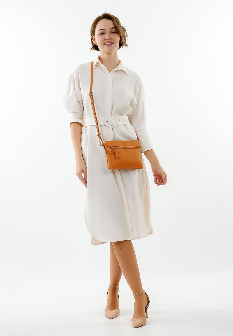 Abito camicia bianco con cintura in vita; abbinato a una piccola borsa a tracolla marrone e tacchi nudi su uno sfondo neutro.