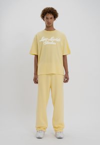 Jeune personne debout, portant un t-shirt jaune surdimensionné avec "Low Lights Studios" et un pantalon ample assorti, accompagné de baskets blanches.