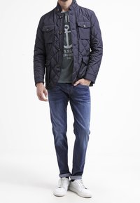 Quilted navy jacket with a high collar, two front pockets, and button detailing, worn over a grey t-shirt and blue jeans with white sneakers.