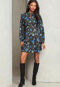 Schwarzes Blumenkleid mit blauen Rosen und grünen Blättern, langen Ärmeln und einem Schlüsselloch-Ausschnitt. Kombiniert mit schwarzen kniehohen Stiefeln.