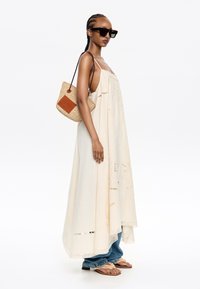 Vestido maxi beige claro con detalles bordados, combinado con jeans azules y sandalias color beige. Accesorio: bolsa de mimbre con acento de cuero. Gafas de sol negras.