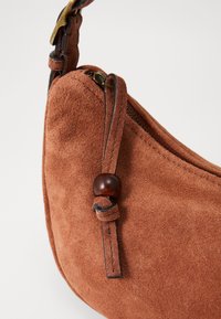 Borsa a spalla in suede marrone con forma arrotondata, dotata di chiusura a zip e un dettaglio decorativo a nodo con un accento in legno.