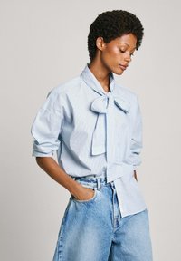 Hellblau gestreiftes Hemd mit einem großen Fliegenkragen, hochgekrempelten Ärmeln und einer Hose aus hellblauem Denim mit hoher Taille.
