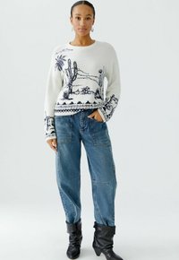 Witte trui met een desertlandschap ontwerp in marineblauw, gecombineerd met blauwe baggy jeans en zwarte kniehogere laarzen. Geweven brei stof.