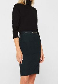 Schwarzer Rollkragenpullover und dunkelgrüner Bleistiftrock mit seitlichem Schlitz, versehen mit einem schmalen schwarzen Gürtel und einem schlichten, klaren Design. Weicher, strukturierter Stoff.