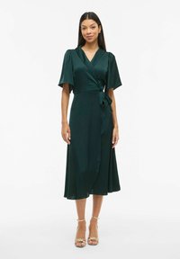 Omslagskjole i dyp grønn sateng, med korte puffermede ermer, et knyttet midjebelte og en flytende A-linje silhuett, ankel-lang.