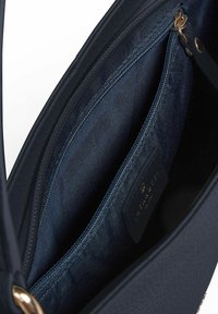 Innenraum einer Marine-Handtasche mit glatter Textur, Reißverschlusstasche und einem Marke-Label. Ausgelegt mit dunklem Stoff, der einfache Designelemente präsentiert.