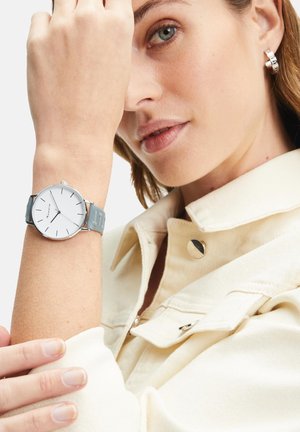 Montre argentée avec un cadran blanc et un bracelet en silicone gris. La personne qui la porte a également une boucle d'oreille en argent et est accompagnée d'une veste crème claire.