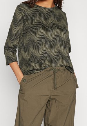 Personne portant un haut rayé ondulé vert olive et noir à manches trois quarts et un pantalon vert olive à cordon avec des poches boutonnées.